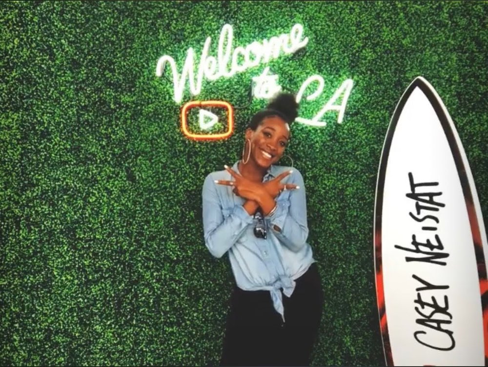 Ada smiles and holds up the peace sign in front of a neon sign reading “Welcome to LA” and a surfboard that reads “Casey Neistat”