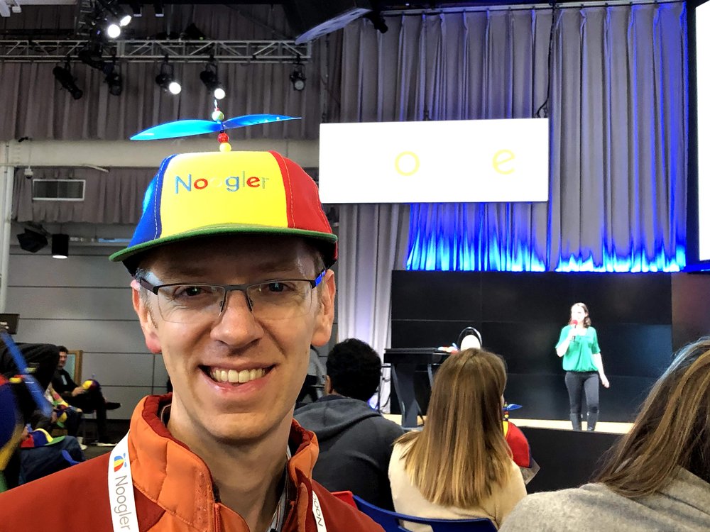 Garrett wearing a Noogler hat. In the background are other people sitting in chairs and a person standing on a stage holding the microphone.