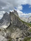 A photograph of a mountain landscape, with a dramatically slanted mountain in the foreground that reveals a green pasture on one side and a rocky wall on the other.