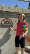 A woman in a red t-shirt holding a colourful windmill pointing at a plaque of a rainbow, with a sign underneath that says “Coogee Rainbow Walkway. Diversity, Inclusion, Love”