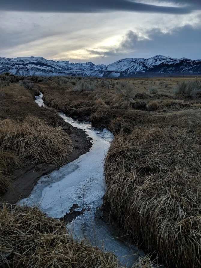 Photo showing a river with fields on either side of it leading to a mountain range in the foreground.