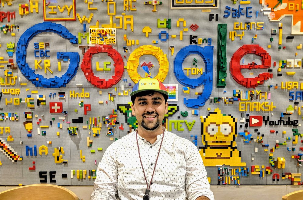 Nikhil wearing a Noogler hat sitting in front of a wall made of legos.