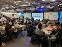 A group of people sit in a cafeteria at dining tables, eating together. Screens at the head of the room read: “Ramadan Kareem” and “Google.”