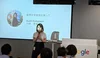 Ikumi speaking into a microphone in front of a large group. A slide is projected behind her introducing herself.