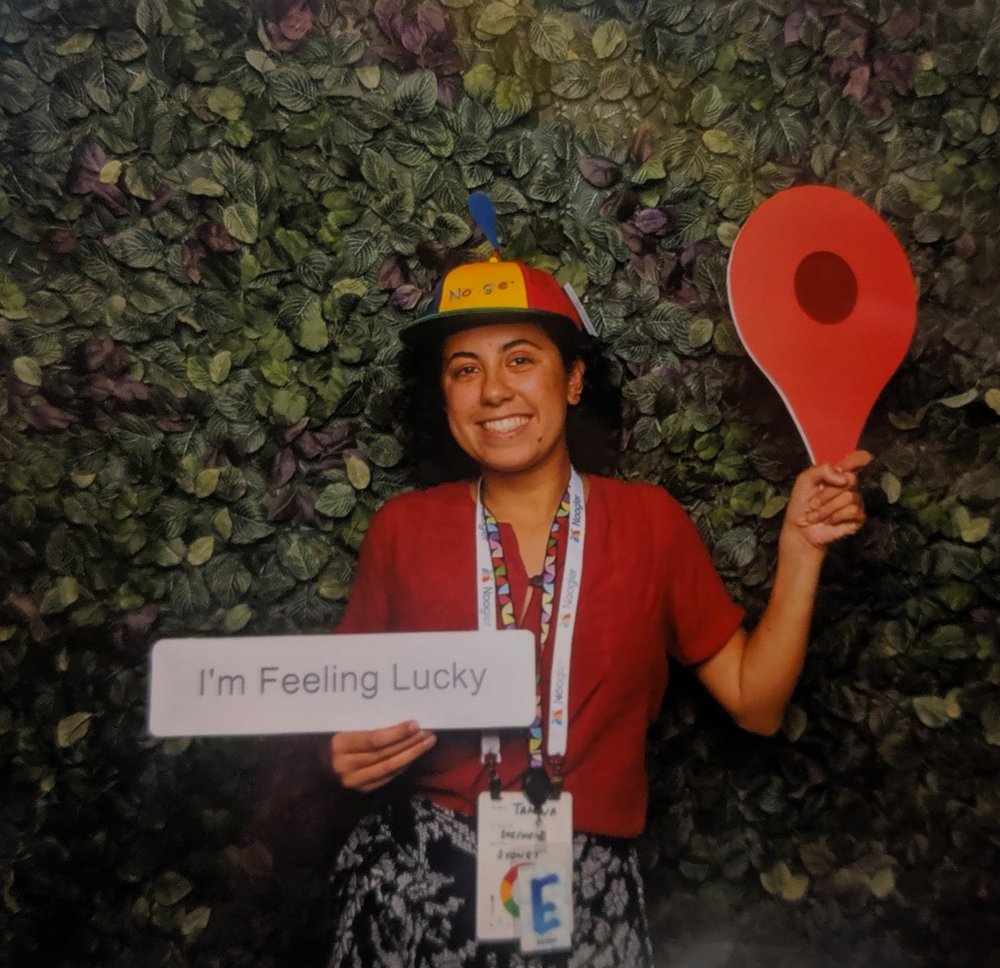 Tamina stands in front of a wall of leaves. She is wearing a red shirt with a black-and-white floral skirt. She holds cardboard signs of the Google Maps red logo and the Google “I’m Feeling Lucky” search button. She also wears a Noogler hat — a green, blue, red and yellow hat with a propeller.