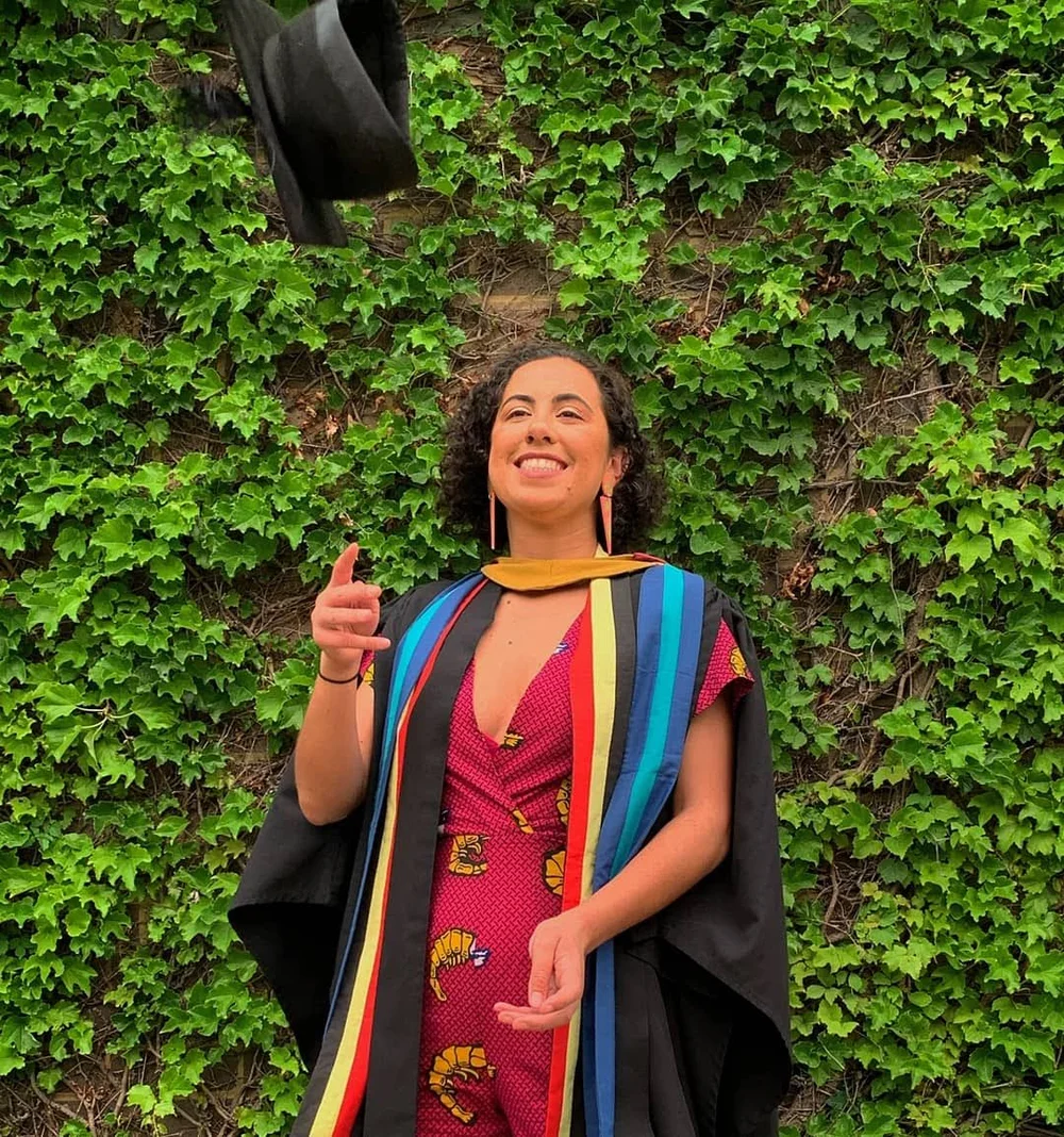 Tamina stands outdoors in front of a wall of greenery, tossing a graduation cap. She is wearing a red outfit with a shrimp pattern, a black graduation robe, a red, yellow and black sash (the colors of the Aboriginal flag) and a blue and green sash (the colors of the Torres Strait Islander flag).