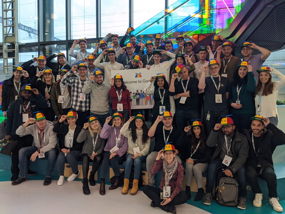 A group of new Googlers (“Nooglers”) wearing colorful propeller caps pose around a “Welcome to Google” banner. In the background are large glass windows looking out to a blue sky.