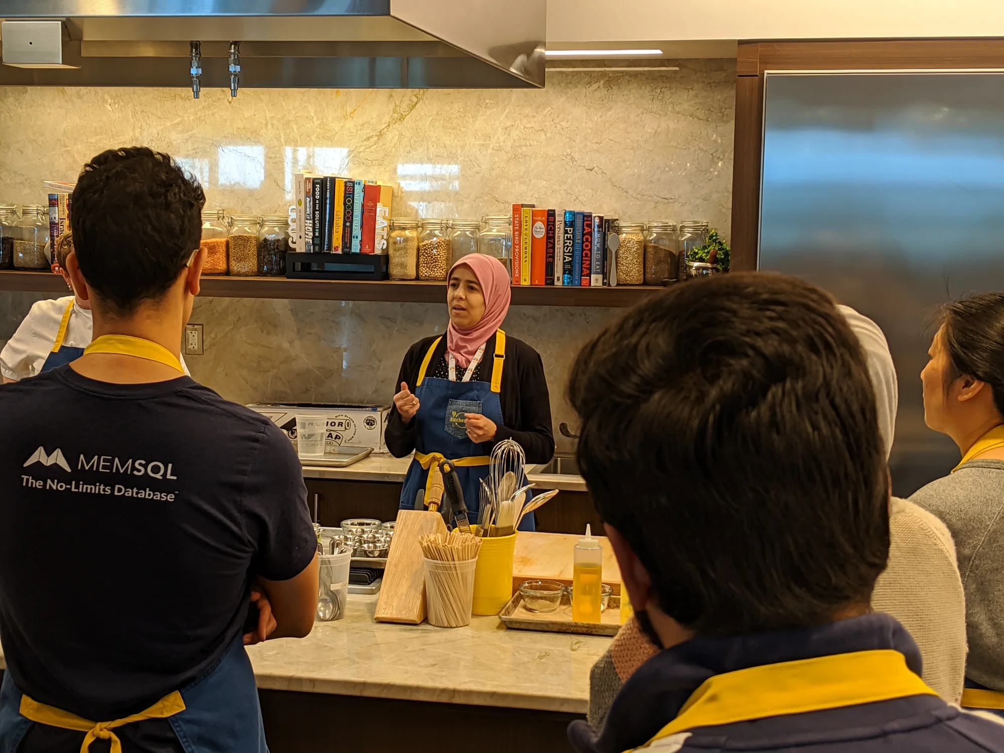 A person wearing an apron stands in front of a group, speaking. The person is behind a table with cooking utensils on it. Everyone wears an apron