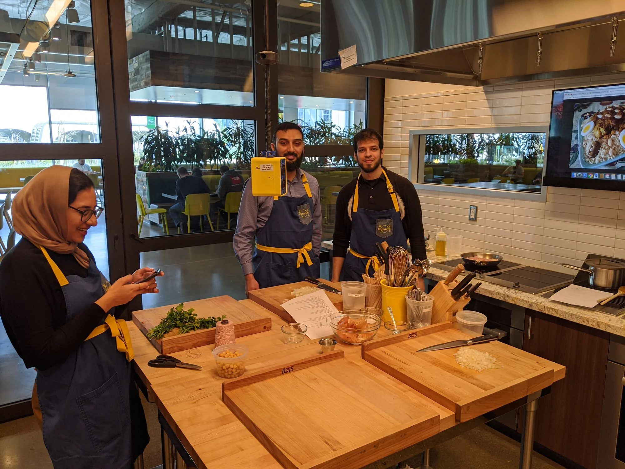 Three people wearing aprons stand near a table with ingredients and cooking utensils on it.