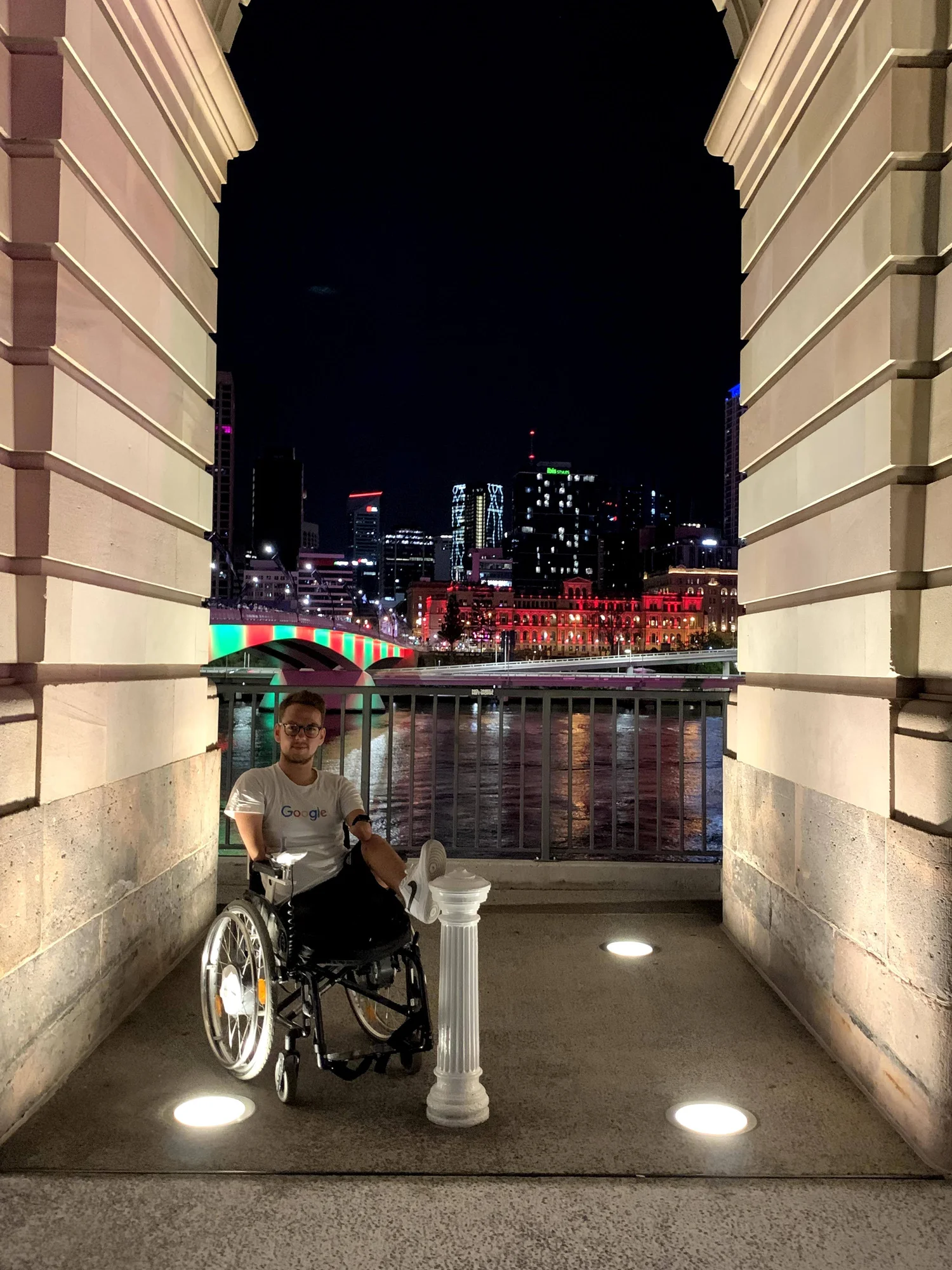 Ein Foto von Patrick Schilling im Rollstuhl sitzend. Im Hintergrund ist die nächtliche Skyline von Brisbane, Australien zu sehen.