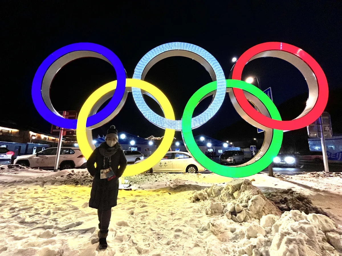 A female Google employee in winter wear, standing in front of the Winter Games logo in Beijing, China.