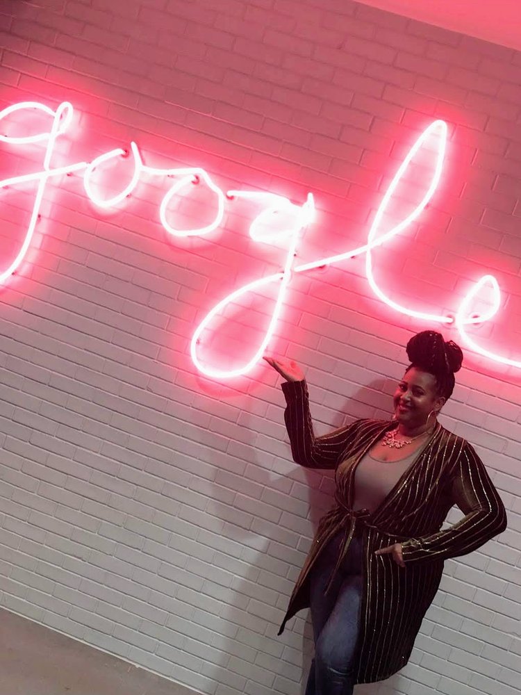 Jennifer stands next to and gestures towards a neon red cursive Google sign