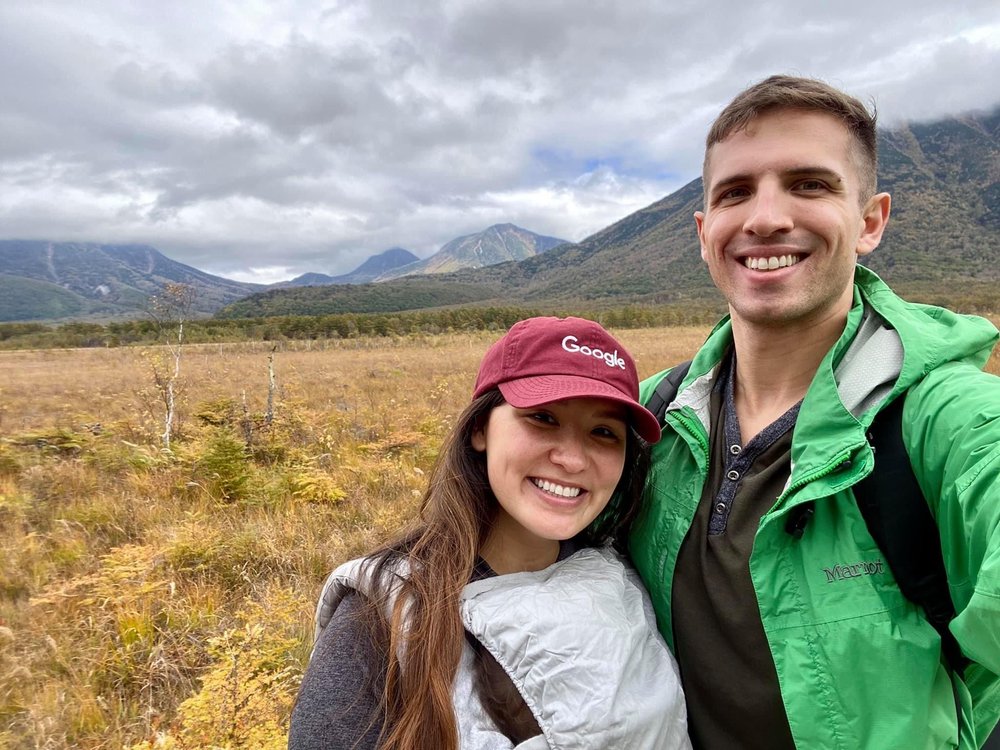 Bry, with a baby in a baby carrier, poses with her husband in front of mountains.