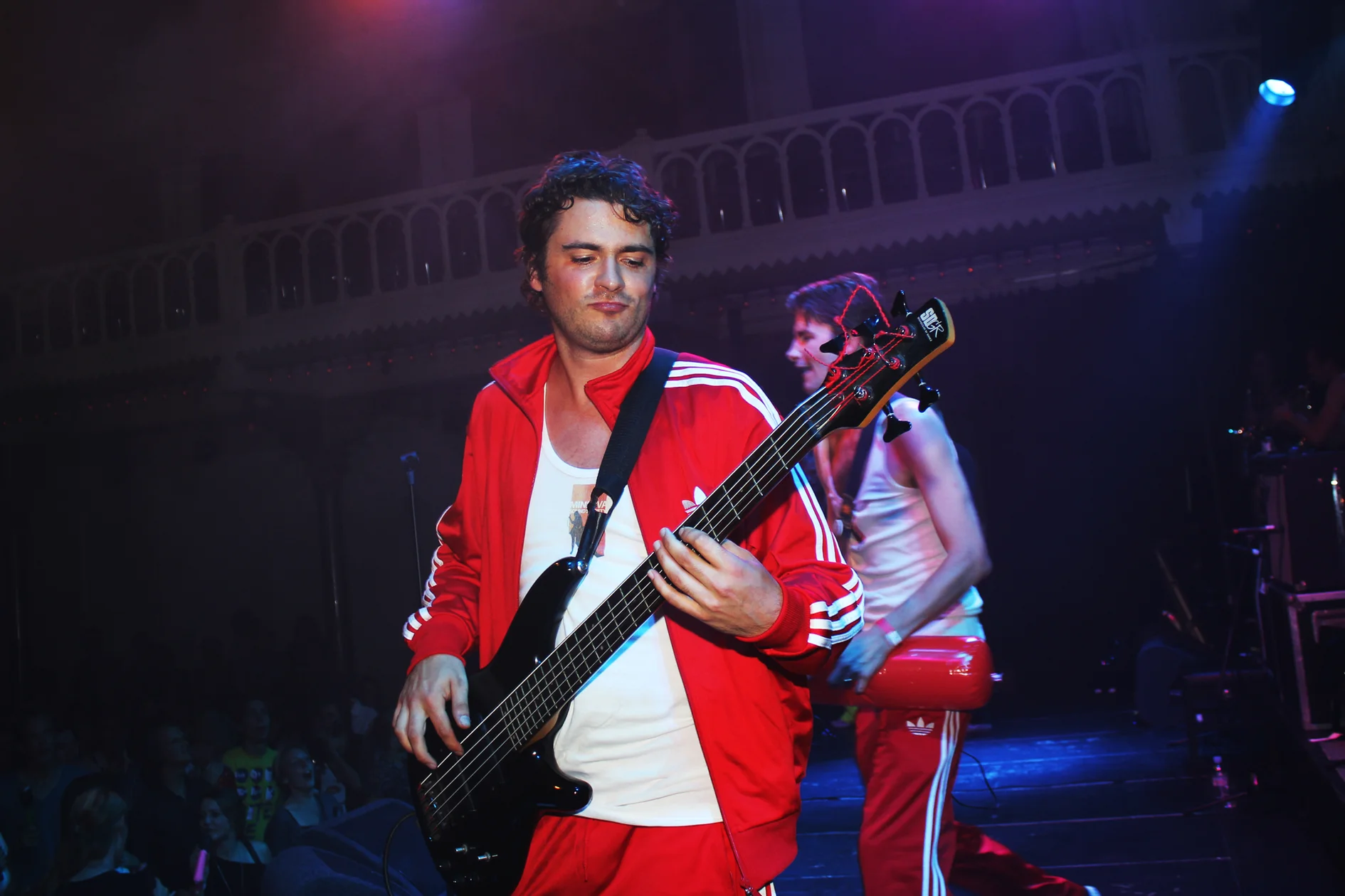 Ben performing on stage. He is playing a bass and wearing a red and white track suit.