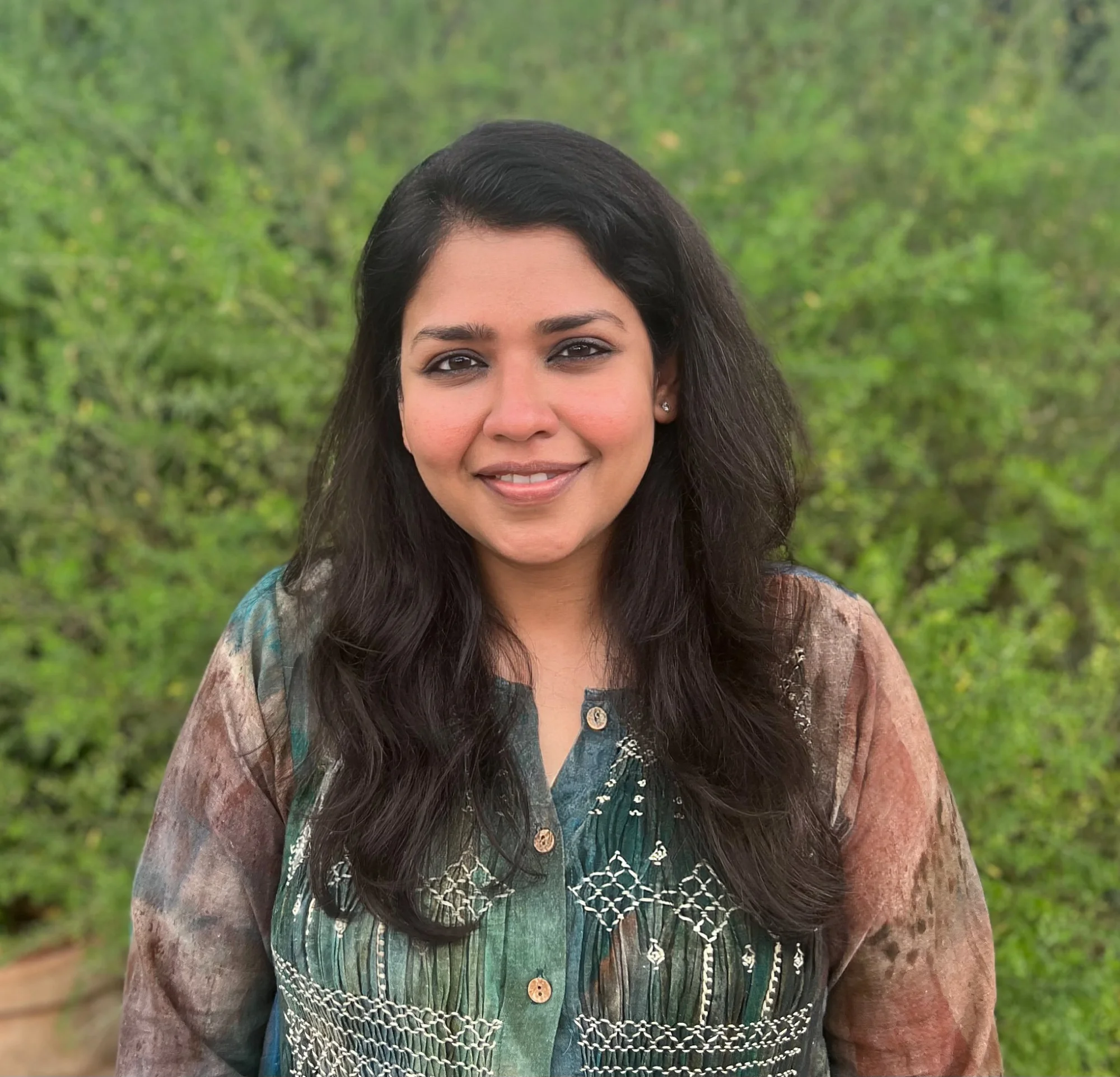 A headshot of a person with long dark hair, a dark green top, against a backdrop of plants