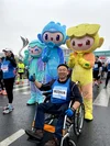 Male Googler in a wheelchair posing for the camera with a thumbs up. He is at a running marathon and is wearing his running attire with a race bib. Behind him are three colorful mascots from the marathon organizer.