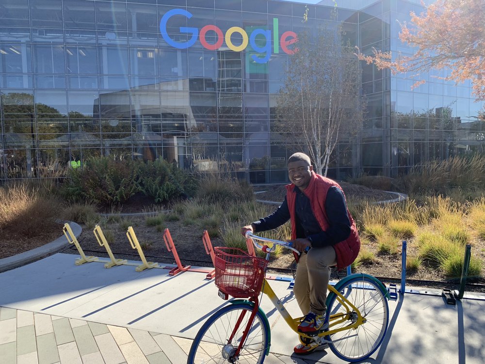 Rob outdoors on a red, yellow, blue, and green “Google bike” in front of the large Google sign at Google headquarters in Mountain View, California.