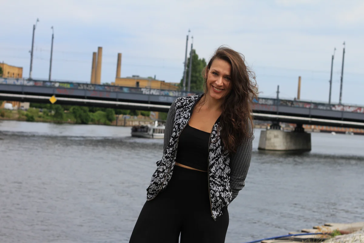 Saskia Bobinska, a woman with long brown hair, stands smiling in front of a river and bridge