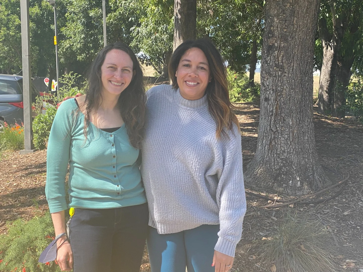 Two women standing side by side with their arms around each other, looking into the camera and smiling. They are standing in front of shrubs and trees, standing on a sidewalk next to yellow and blue circular objects on the sidewalk.