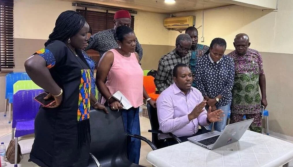 A man in front of a laptop is tutoring 8 others who are standing behind him and  staring at the laptop