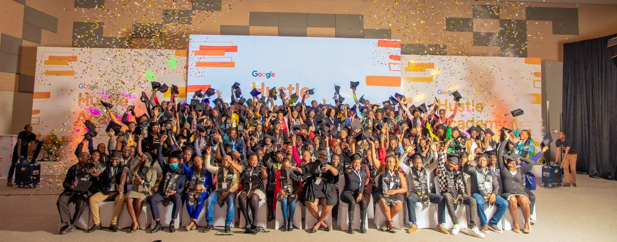 A group of joyful graduates at the Google Hustle Academy event, tossing caps among confetti. Hustle Academy branding is visible in the background.