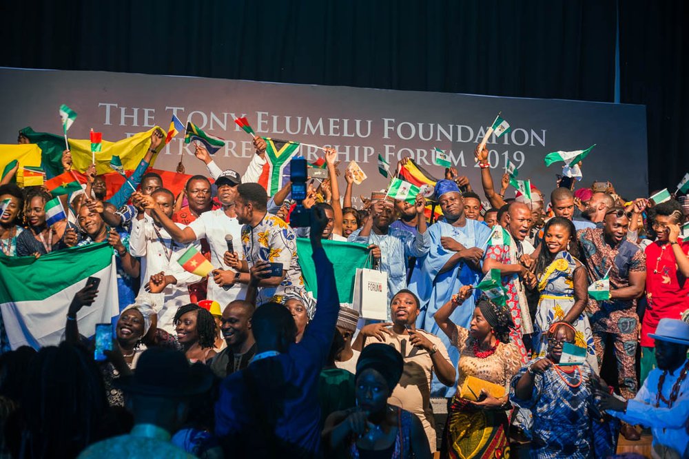 A crowd of African entrepreneurs are celebrating up on a stage with a backdrop sign for the Tony Elumelu Foundation