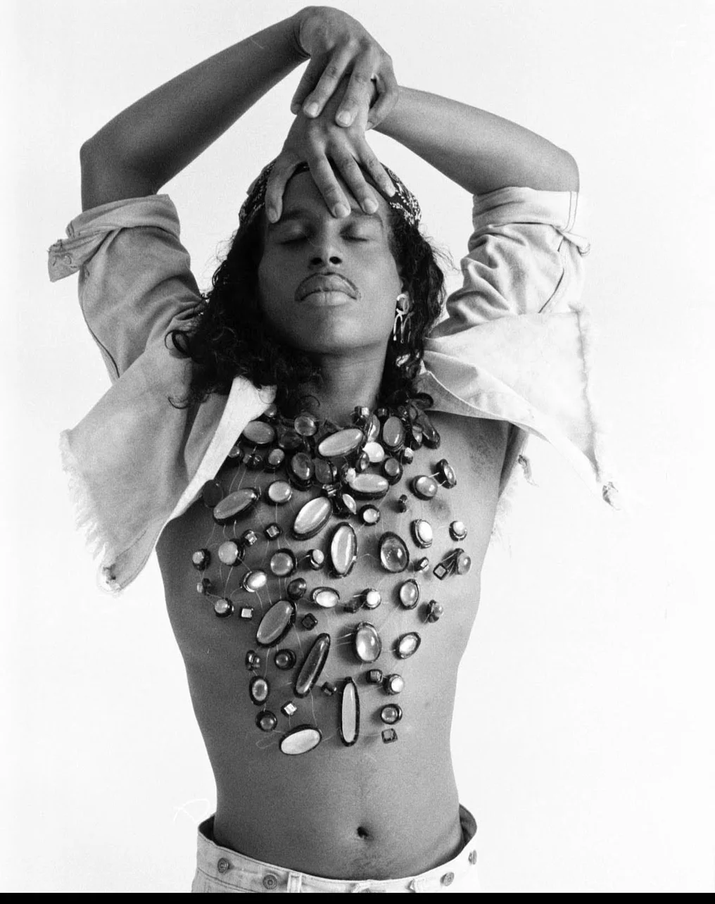 Willi Ninja vogues for the camera, with his arms and hands above his head. He looks upward with his eyes closed, appearing reverant as his bare torso is adorned in an intricate bodysuit by French fashion designer Thierry Mugler made of large glass beads.