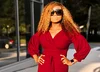 Wearing a long-sleeved red dress, big sunglasses and hoop earrings, Tokes poses outside of an office building.