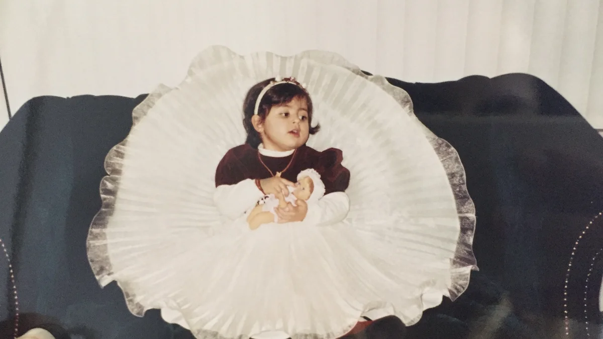 The author as a child, dressed up for Eid. She is holding a doll and wearing a fancy dress with a full skirt that is fanned out around her.