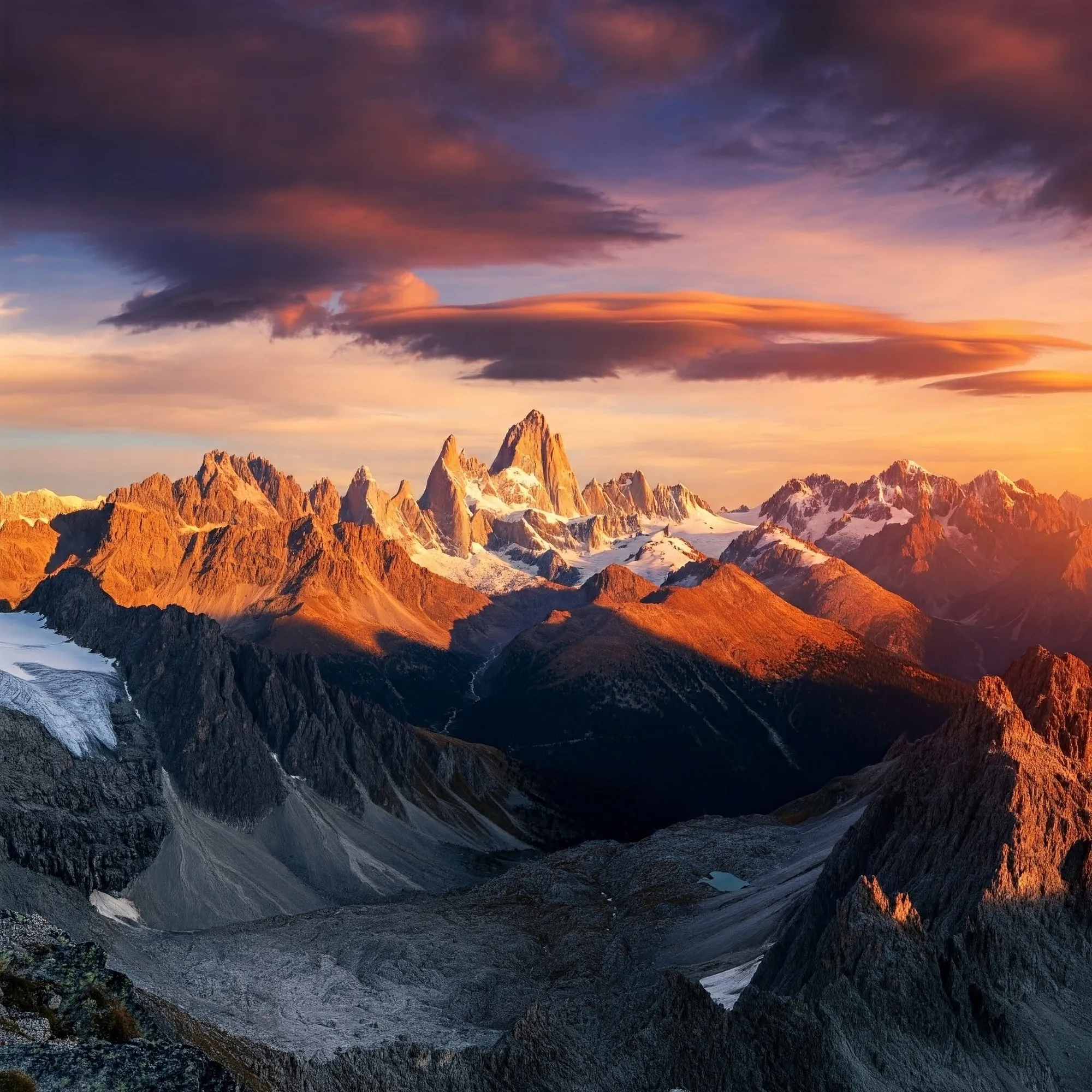 Una imagen fotorrealista de una impresionante vista de montaña con picos escarpados y cumbres nevadas, bañadas por el cálido resplandor de un sol poniente. Dramáticas nubes, pintadas en vibrantes tonos naranja, rosa y morado, surcan el cielo proyectando largas sombras sobre el escarpado paisaje.