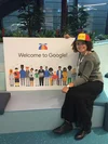 Inna Khlon sitzt auf einer Bank, trägt eine bunte Noogler-Mütze und hält ein Schild in die Kamera, auf dem „Willkommen bei Google“ in englischer Sprache zusammen mit einigen Illustrationen von verschiedenen Menschen abgebildet ist.