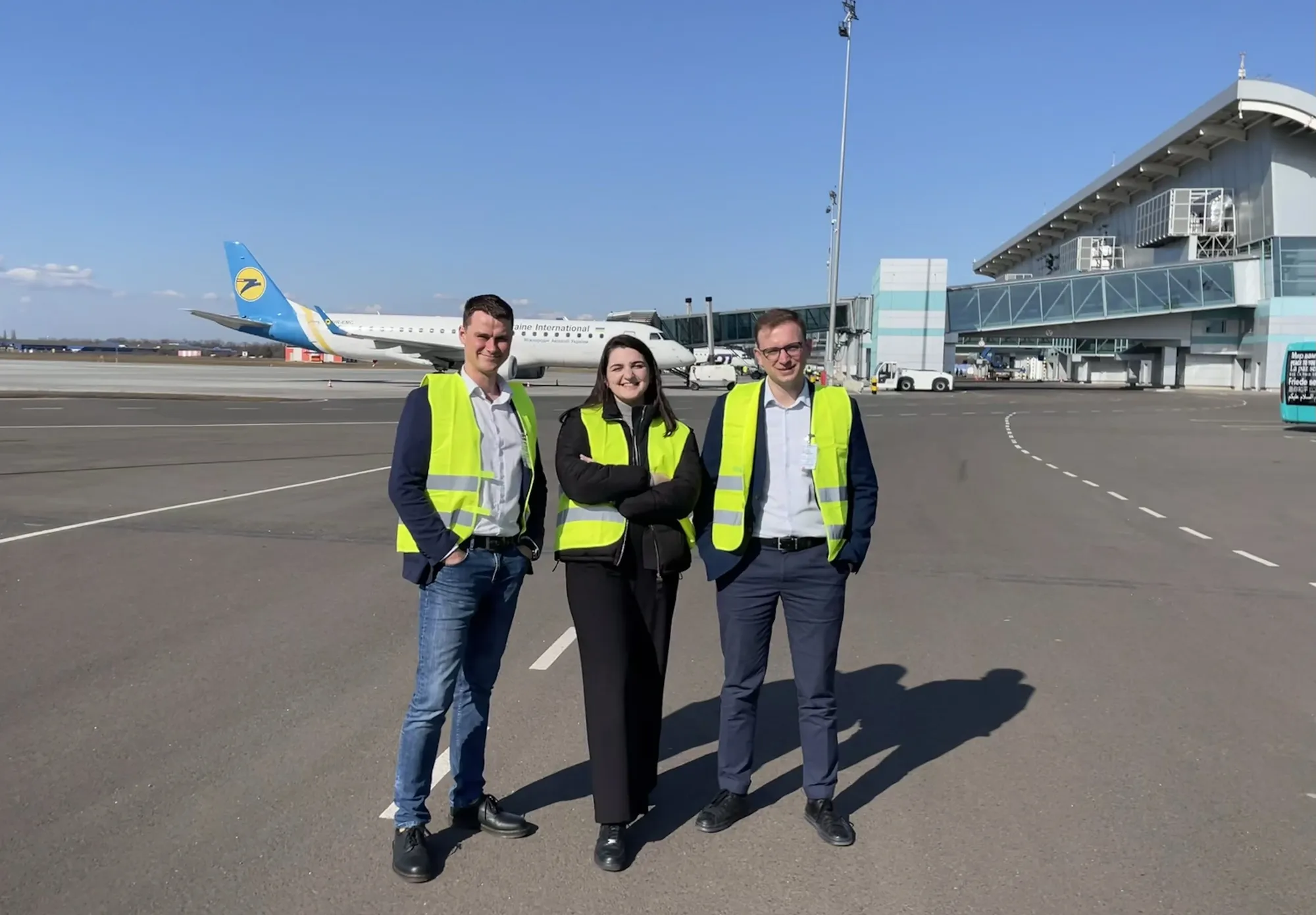 A woman and two men stand on the airfield in front of a plane.