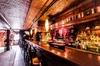 Interior photograph of Stonewall Inn bar without people inside. A wooden bar and stools are visible and alcohol is lined up behind the bar, along with T-shirts for sale.