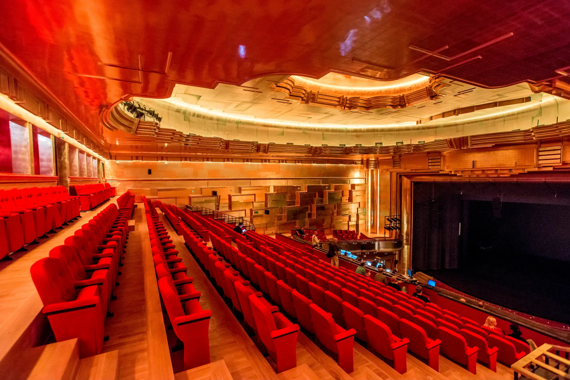 The image shows the interior of the Capitol Musical Theater, a modern and luxurious performance venue. The theater features rows of bright red, plush seats arranged in ascending tiers. The walls and ceiling are decorated with warm shades of gold and copper, featuring textured geometric patterns that add depth and sophistication, while soft, strategically placed lighting highlights the architectural details and creates a cozy atmosphere. The large stage, framed by golden columns and intricate paneling, complements the elegant design, making the theater an inviting and visually appealing space.