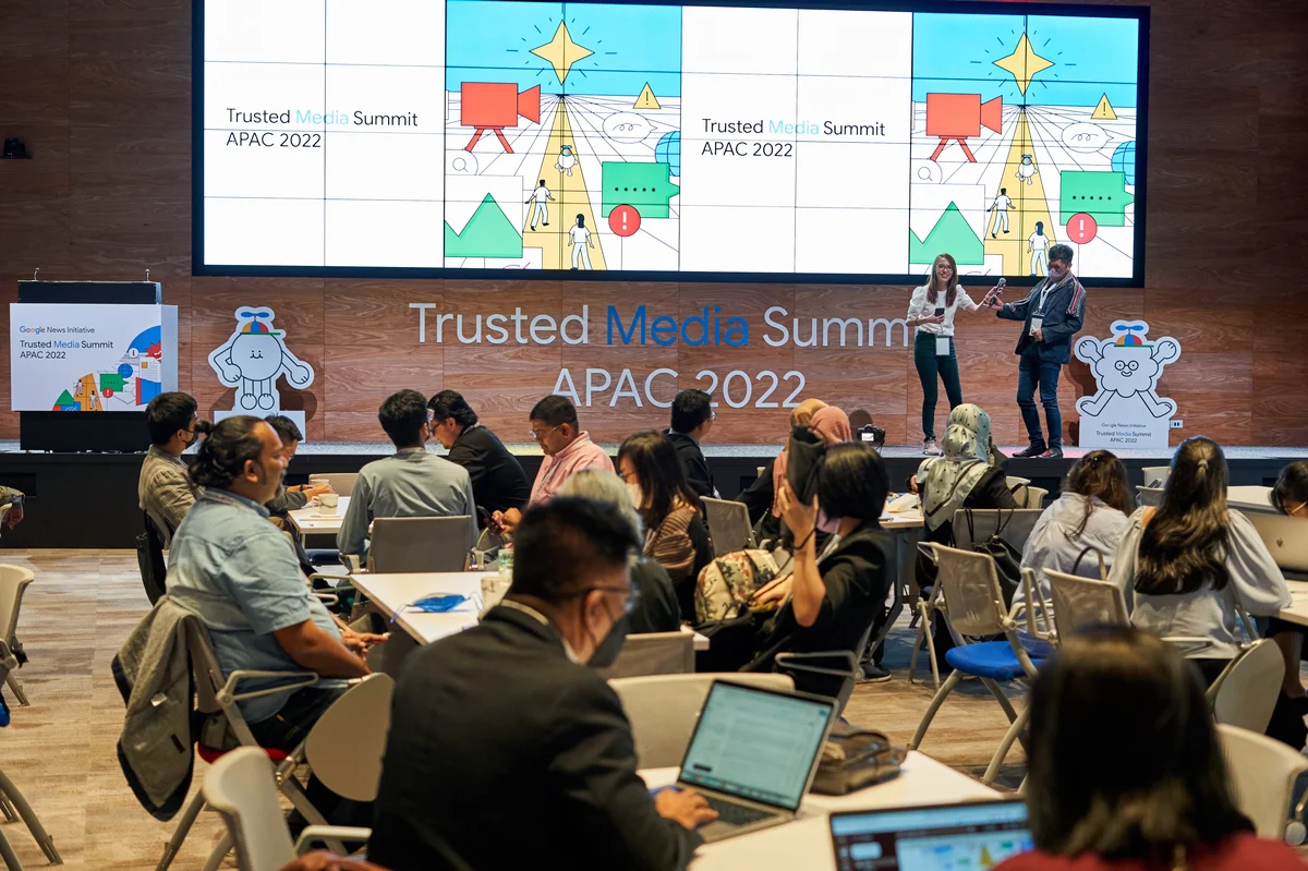 Group of people sitting at tables with laptops, looking at a stage labeled "Trusted Media Summit APAC 2022".