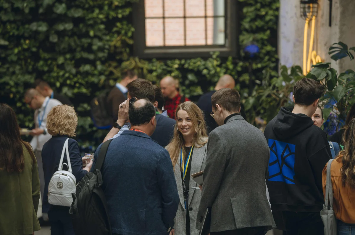 A group of people speak in an open-air area, with green vines on the walls. A blonde woman wearing a yellow and blue lanyard and a gray blazer is looking at the camera.