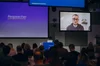 Several screens hang from the ceiling in front of crowd. One the righthand screen, a man wearing glasses and a black sweatshirt looks at the camera. On the lefthand screen, the word "Respeecher" is written in white on top of a blue background. In between the screens is a podium that says "Ukraine Support Fund."