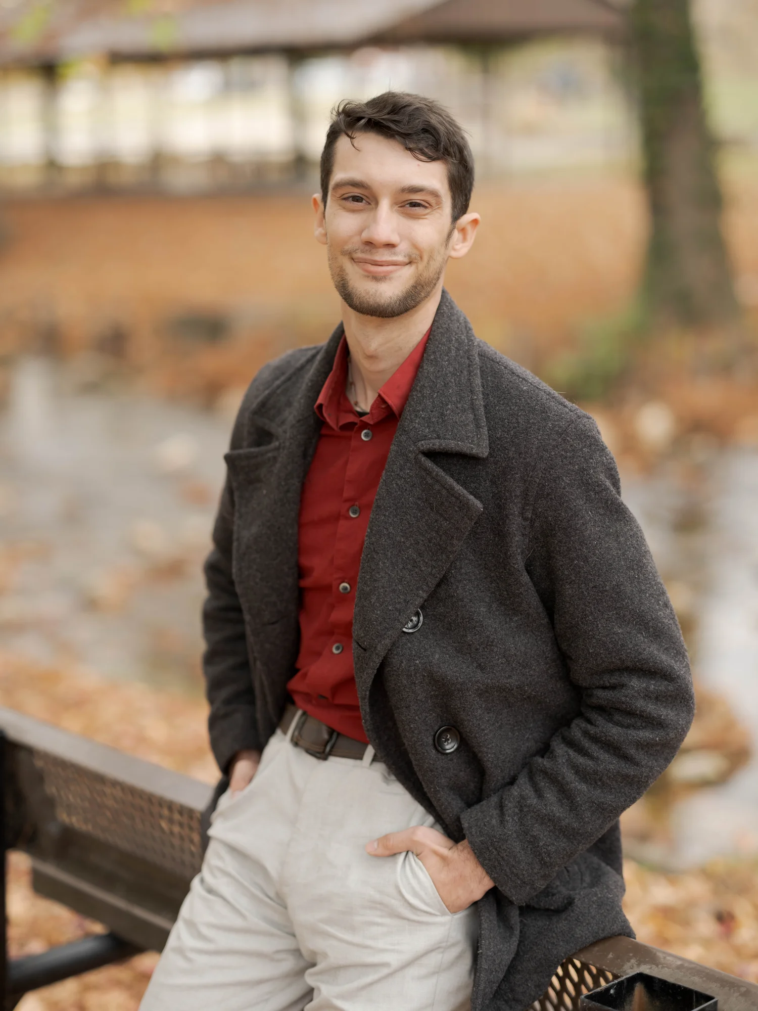 A man wearing a brown blazer smiles directly at the camera as he stands outside in front of a tree.