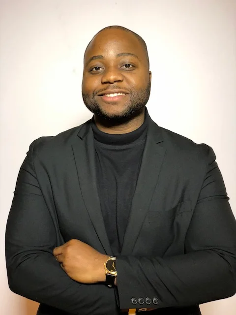 Jeffrey Osuji smiles at the camera with his arms crossed, wearing a black turtleneck, black blazer, and with a black and gold watch on.