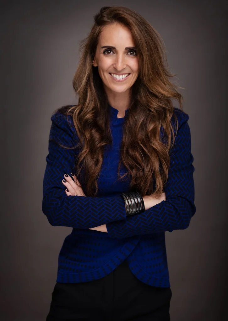 A woman with long brown curly hair and wearing a blue blazer smiles at the camera, with her arms folded across her chest.