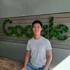 Jiehua Wu, wearing a white t-shirt and blue pants, sits on a wooden bench in front of a green Google logo.