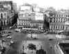 A black-and-white photograph of a city square in the 1950s, with cars driving by the streets and billboards on the building façades