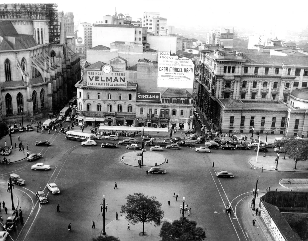 Uma fotografia em preto e branco de uma praça da cidade na década de 1950, com carros passando pelas ruas e outdoors nas fachadas dos edifícios