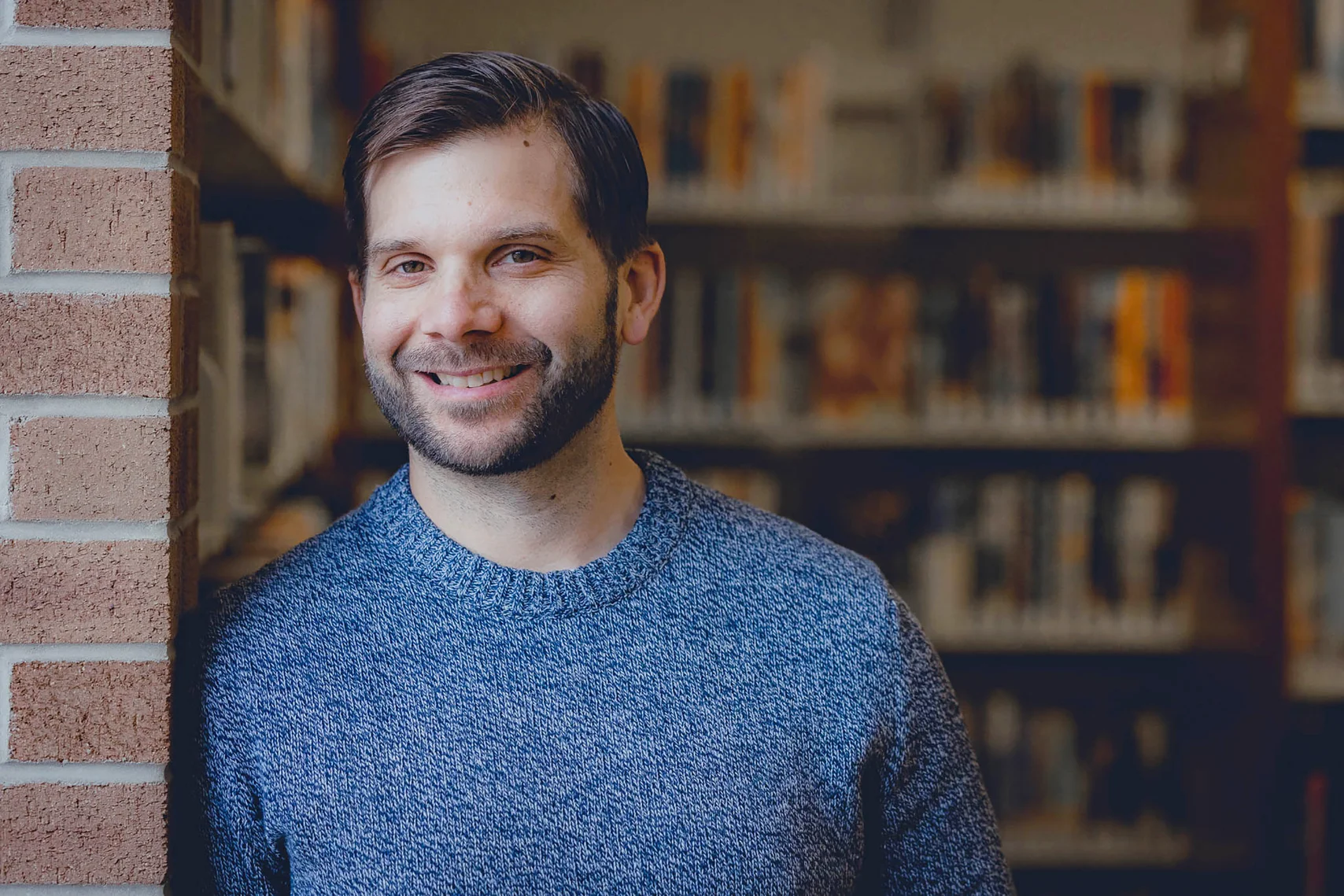 A man wearing a blue sweater leans his shoulder against the wall and smiles directly at the camera.