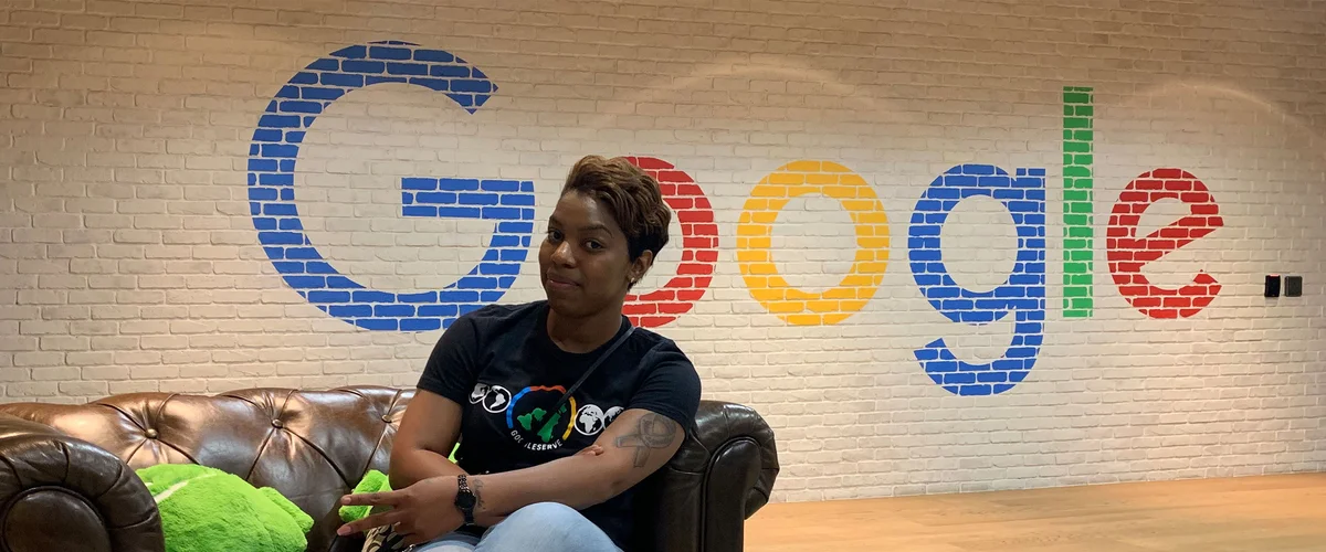 Joy sits on a brown couch. Behind her is the Google logo painted on a white brick wall.