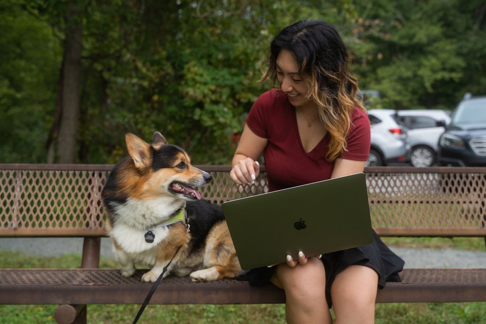 Julia sits on a metal bench with her dog, Keto. She is holding her laptop open and gesturing to the screen.