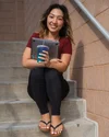 Julia smiles at the camera while sitting on concrete stairs, holding a cup and her laptop.