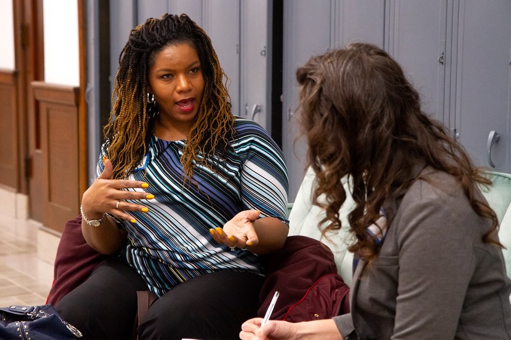 Picture shows two women sitting on a sofa. The woman on the left is speaking and gesticulating with her hands while the woman on the right is listening and taking notes.