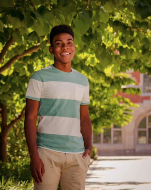 Kahlil stands outside, in front of a tree in a green and white striped shirt and khaki pants. A building can be seen in the distance.