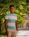 Kahlil stands outside, in front of a tree in a green and white striped shirt and khaki pants. A building can be seen in the distance.